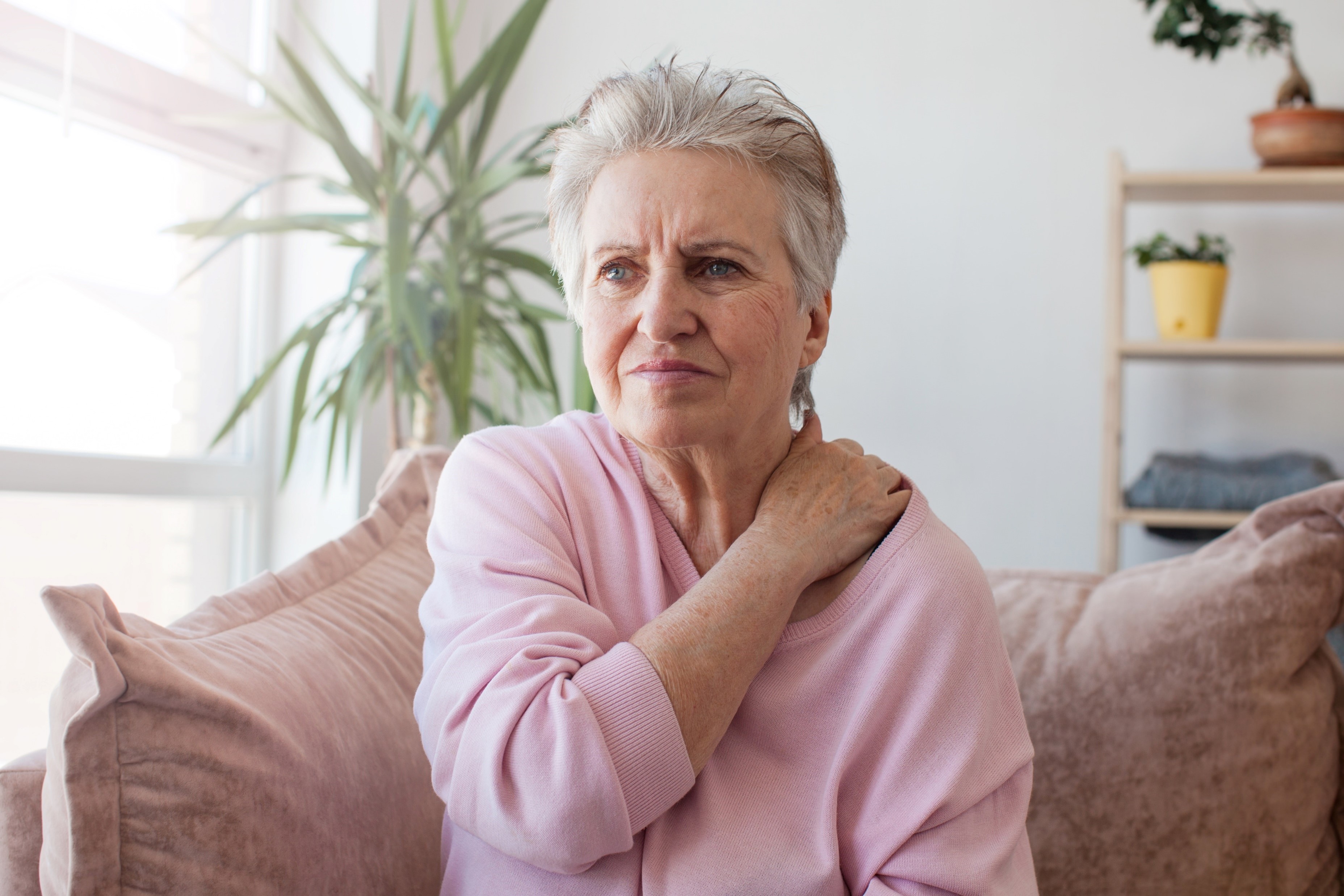 Oudere dame pakt haar linkerschouder vast met haar rechterhand en kijkt of ze pijn heeft.